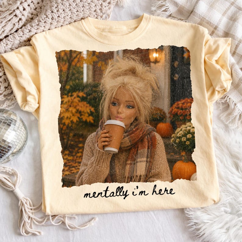 Op de afbeelding: Een lichtgeel t-shirt met een rechthoekige grafische print van een blonde pop met warrig haar, gekleed in een bruine trui en een geruite sjaal, terwijl deze een koffiebeker vasthoudt. De achtergrond toont herfstbladeren en pompoenen. De tekst luidt mentally i'm here.
