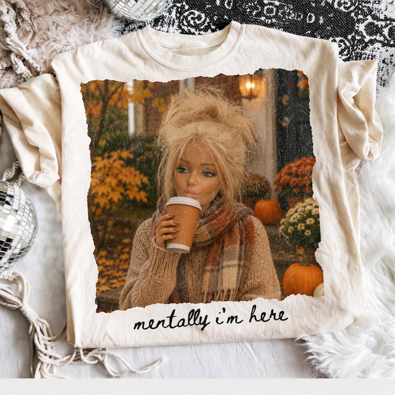 Op de afbeelding: Een cr&egrave;mekleurig t-shirt met een rechthoekige afbeelding van een persoon met blond haar in een bruine trui en sjaal, die een koffiebeker vasthoudt in een herfstsetting met pompoenen. De tekst "mentally i'm here" is in het zwart onder de afbeelding gedrukt.