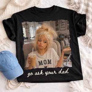 May include: Black t-shirt featuring a distressed image of a woman holding a drink, with the word "MOM" and the phrase "go ask your dad". A blue denim baseball cap is visible on the left.