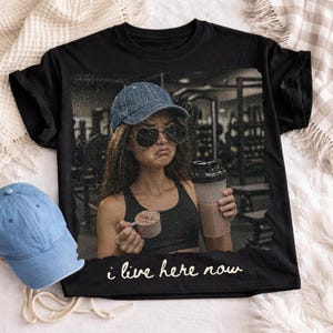 May include: Black t-shirt featuring a distressed image of a person in a gym setting, wearing sunglasses and a denim cap, holding a protein shake and scoop. The text "i live here now" is printed at the bottom.