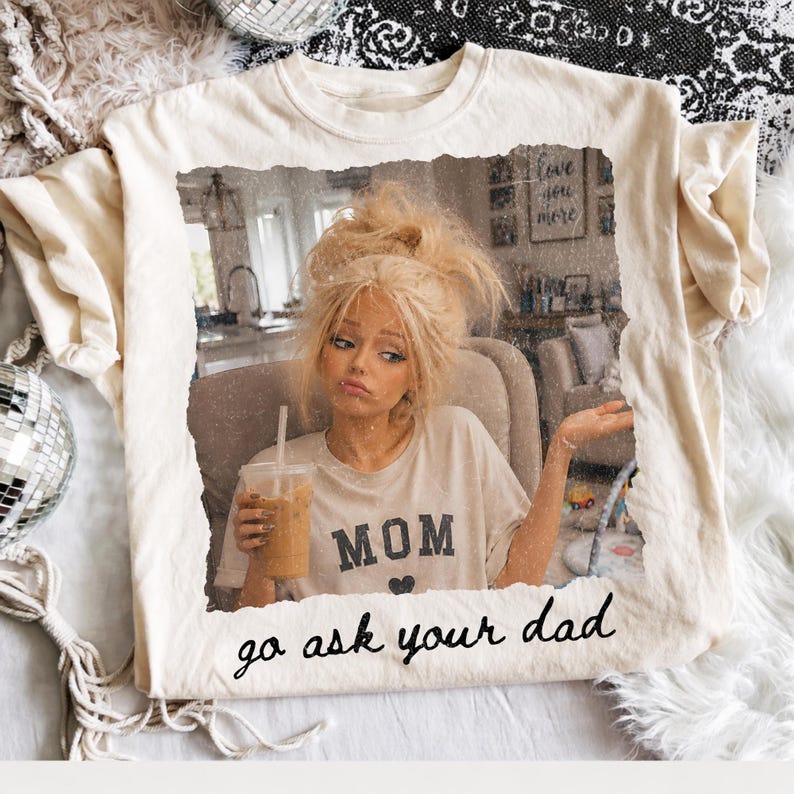 May include: Cream-colored t-shirt featuring a distressed image of a woman holding a drink. The shirt has the word "MOM" with a heart and the phrase "go ask your dad" printed on it. The shirt is on a white surface.
