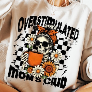 May include: White sweatshirt with a graphic of a skeleton wearing sunglasses and a bandana, holding an orange mug. The text reads "OVERSTIMULATED MOM'S CLUB" with daisy and sunflower accents.