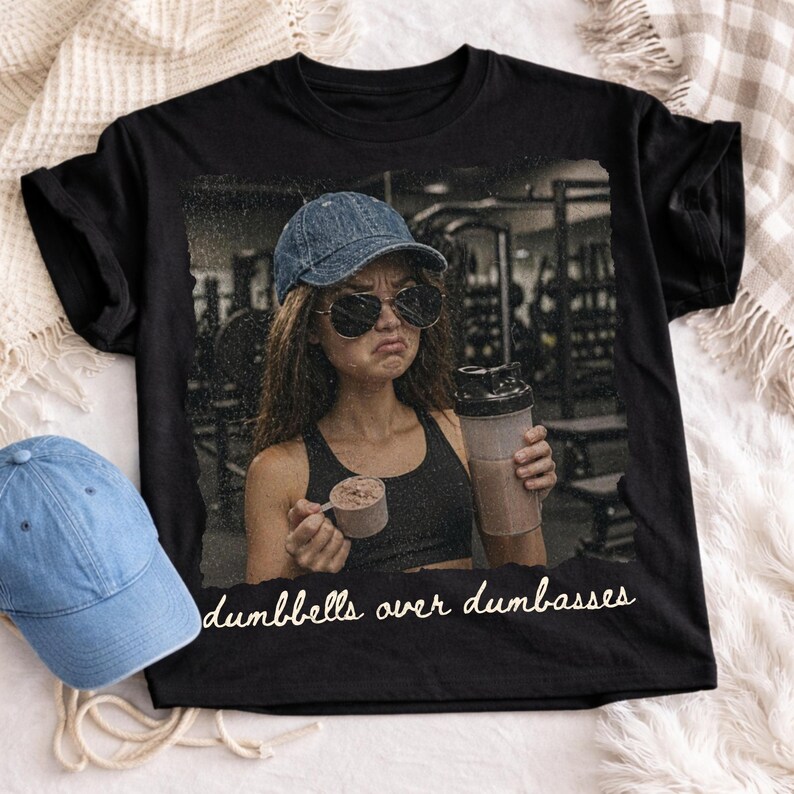 May include: Black t-shirt featuring a distressed image of a person in a gym setting, wearing sunglasses and a denim cap, holding a protein shake and scoop. The text "dumbbells over dumbasses" is printed in white.