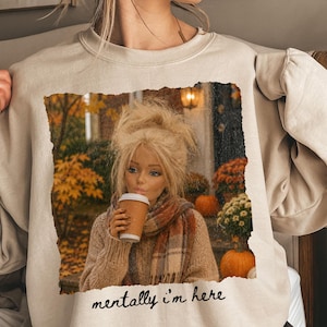 Op de afbeelding: Een beige sweater met ronde hals en een rechthoekige grafische print van een blonde pop in een beige trui en geruite sjaal, terwijl deze een koffiebeker vasthoudt. De achtergrond van de print toont herfstbladeren en pompoenen. Tekst: mentally i'm here.