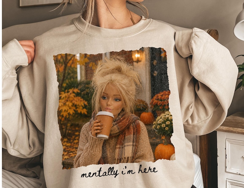 Op de afbeelding: Een beige sweater met ronde hals en een rechthoekige grafische print van een blonde pop in een beige trui en geruite sjaal, terwijl deze een koffiebeker vasthoudt. De achtergrond van de print toont herfstbladeren en pompoenen. Tekst: mentally i'm here.
