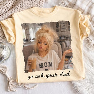 May include: Cream-colored t-shirt featuring a distressed image of a woman holding a drink. The shirt has the word "MOM" with a heart, and the phrase "go ask your dad" in a handwritten font.