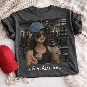 May include: Dark gray t-shirt featuring a distressed image of a person wearing sunglasses and a denim cap, holding a protein shake and scoop. The text "i live here now" is printed on the shirt. A red baseball cap is visible on the left.