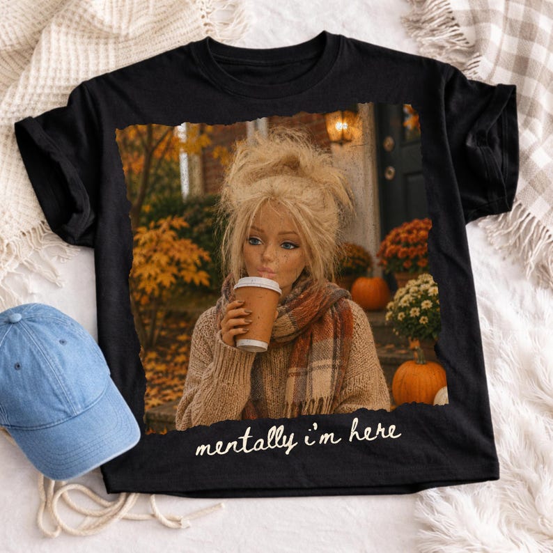 Op de afbeelding: Een zwart t-shirt met een grafische print van een persoon met blond haar in een beige trui en sjaal, die een koffiebeker vasthoudt. De sc&egrave;ne bevat herfstbladeren en pompoenen. De tekst "mentally i'm here" staat in het wit eronder gedrukt.