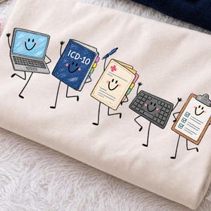 Puede incluir: Una camiseta de color crema con un estampado gráfico de cinco artículos de oficina antropomórficos con caras sonrientes: un portátil, un libro azul ICD-10, un historial médico con una cruz roja, un teclado negro y un portapapeles con lista.