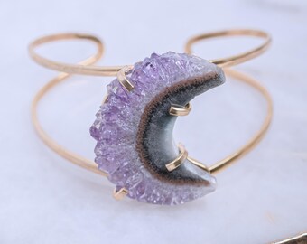 May include: Gold-tone metal cuff bracelet featuring a crescent-shaped amethyst stone. The amethyst has a purple, gray, and brown color scheme. The bracelet has a simple, open design.