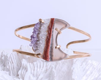 May include: A gold cuff bracelet with a crescent moon shaped agate stone. The stone is layered with amethyst crystals, white quartz, and red agate.