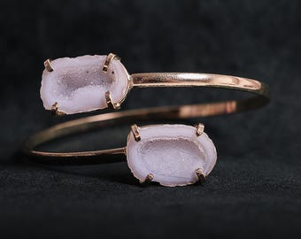 May include: A gold-coloured cuff bracelet with two oval, pale purple geode stones. The stones have a crystalline interior and are set in a gold metal frame. The bracelet has a simple, elegant design, suitable for everyday wear.
