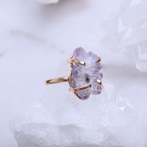 May include: A gold ring featuring a large amethyst crystal cluster. The crystal is a light purple colour and has a natural, rough texture. The ring is set against a white background.