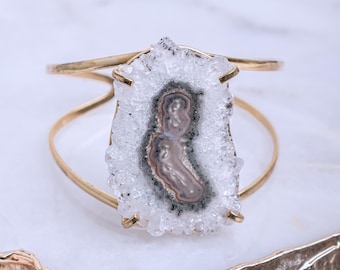 May include: A gold metal cuff bracelet with a large, clear quartz crystal featuring a gray and white agate pattern.