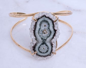 May include: A gold cuff bracelet with a large, green and white agate stone set in the center. The stone has a unique, swirling pattern and is surrounded by clear quartz crystals.