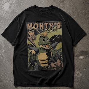 Puede incluir: Camiseta negra con un cocodrilo de dibujos animados con gafas de sol y cresta, sosteniendo un palo de golf. La camiseta tiene el texto "Monty's Gator Golf" en un estilo vintage. El diseño está impreso en una tela oscura y descolorida.
