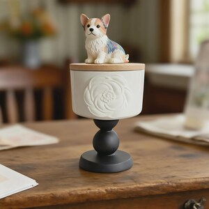 Peut inclure: Un r&eacute;cipient d&eacute;coratif en c&eacute;ramique blanche avec un couvercle en bois, orn&eacute; d'un motif floral sculpt&eacute;. Une petite figurine de corgi color&eacute;e est pos&eacute;e sur le couvercle. Le r&eacute;cipient est soutenu par un socle noir, cr&eacute;ant un pr&eacute;sentoir &agrave; plusieurs niveaux.