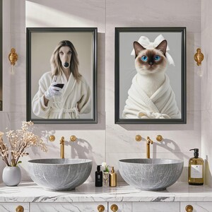 May include: Two framed art prints. One features a dog with a human body wearing a white robe and holding a wine glass. The other print shows a cat in a white robe with a white bow. The prints are above two gray marble sinks.