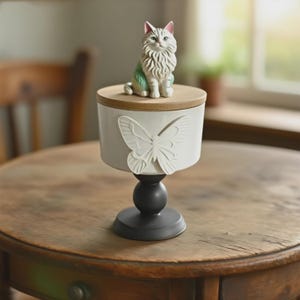 Peut inclure: Une figurine d&eacute;corative de chat blanc et gris est pos&eacute;e sur un pot blanc avec un couvercle en bois. Le pot pr&eacute;sente un motif de papillon en relief et repose sur une base de pi&eacute;destal noire. Le pot est expos&eacute; sur une table en bois.