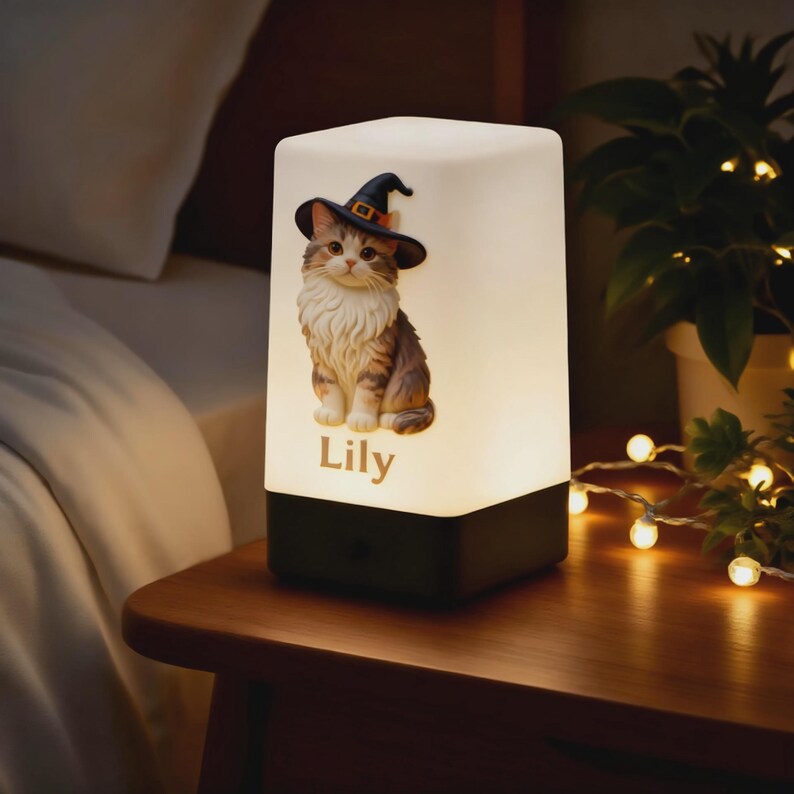 May include: A decorative night light featuring a detailed illustration of a cat wearing a witch hat. The cat is brown and white, with the name "Lily" printed below. The lamp has a black base and a white, glowing top.