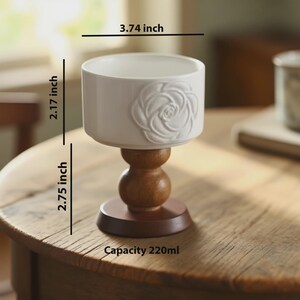 Peut inclure: Une tasse en c&eacute;ramique blanche avec un motif de rose, de 9,5 cm de large, 5,5 cm de haut, et une base en bois de 7 cm de haut. La tasse a une capacit&eacute; de 220 ml. La tasse est sur une table en bois.