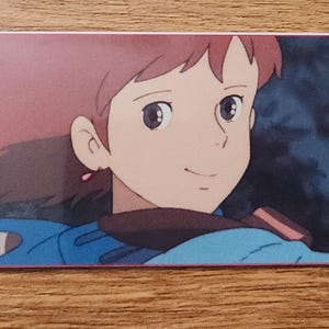 Peut inclure: Un marque-page rectangulaire en papier avec une illustration imprim&eacute;e d'un personnage aux cheveux courts brun-roux, portant une veste bleue et des gants bleus, une main lev&eacute;e en signe de pouce lev&eacute;. Un petit trou est perc&eacute; sur le c&ocirc;t&eacute; gauche.