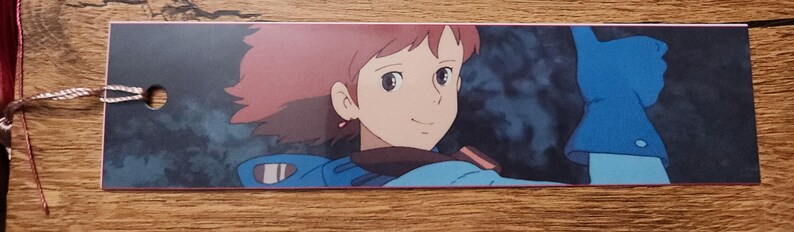 Peut inclure: Un marque-page rectangulaire en papier avec une illustration imprim&eacute;e d'un personnage aux cheveux courts brun-roux, portant une veste bleue et des gants bleus, une main lev&eacute;e en signe de pouce lev&eacute;. Un petit trou est perc&eacute; sur le c&ocirc;t&eacute; gauche.