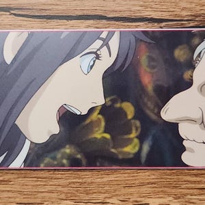 Peut inclure: Un marque-page rectangulaire en papier pr&eacute;sentant une illustration color&eacute;e de style anime d'une jeune personne aux cheveux fonc&eacute;s face &agrave; une personne &acirc;g&eacute;e aux cheveux blancs. Le marque-page a un petit trou circulaire avec un cordon bleu clair.