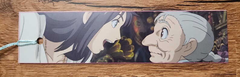 Peut inclure: Un marque-page rectangulaire en papier pr&eacute;sentant une illustration color&eacute;e de style anime d'une jeune personne aux cheveux fonc&eacute;s face &agrave; une personne &acirc;g&eacute;e aux cheveux blancs. Le marque-page a un petit trou circulaire avec un cordon bleu clair.
