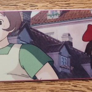 Peut inclure: Un marque-page rectangulaire en papier pr&eacute;sentant une illustration color&eacute;e de style anime d'une personne aux cheveux bruns courts portant une chemise verte et un tablier, tenant une sucette, &agrave; c&ocirc;t&eacute; d'une fille aux cheveux noirs et un grand n&oelig;ud rouge.