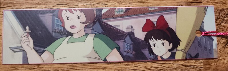 Peut inclure: Un marque-page rectangulaire en papier pr&eacute;sentant une illustration color&eacute;e de style anime d'une personne aux cheveux bruns courts portant une chemise verte et un tablier, tenant une sucette, &agrave; c&ocirc;t&eacute; d'une fille aux cheveux noirs et un grand n&oelig;ud rouge.