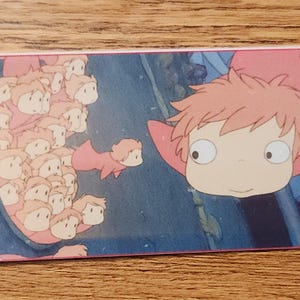 Peut inclure: Un marque-page rectangulaire en papier pr&eacute;sentant une illustration color&eacute;e d'un petit personnage aux cheveux roses et aux grands yeux, accompagn&eacute; d'un groupe de petites figures identiques. Un trou sur la gauche retient un fin cordon rouge et blanc.