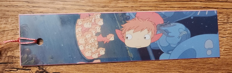 Peut inclure: Un marque-page rectangulaire en papier pr&eacute;sentant une illustration color&eacute;e d'un petit personnage aux cheveux roses et aux grands yeux, accompagn&eacute; d'un groupe de petites figures identiques. Un trou sur la gauche retient un fin cordon rouge et blanc.