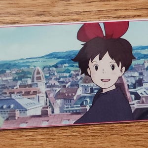 Peut inclure: Un marque-page rectangulaire en papier pr&eacute;sentant une illustration color&eacute;e d'une jeune personne aux cheveux courts fonc&eacute;s et un grand n&oelig;ud rouge, sur un fond de paysage urbain d&eacute;taill&eacute; avec des b&acirc;timents et des toits, fini par un cordon noir.