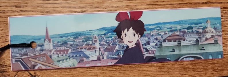 Peut inclure: Un marque-page rectangulaire en papier pr&eacute;sentant une illustration color&eacute;e d'une jeune personne aux cheveux courts fonc&eacute;s et un grand n&oelig;ud rouge, sur un fond de paysage urbain d&eacute;taill&eacute; avec des b&acirc;timents et des toits, fini par un cordon noir.