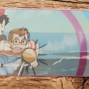 Peut inclure: Un marque-page rectangulaire pr&eacute;sentant une illustration color&eacute;e de style anime de deux personnages faisant du v&eacute;lo ensemble. La sc&egrave;ne montre un arri&egrave;re-plan d'oc&eacute;an bleu avec une rambarde de jet&eacute;e &agrave; gauche et un petit trou avec une ficelle sombre &agrave; droite.