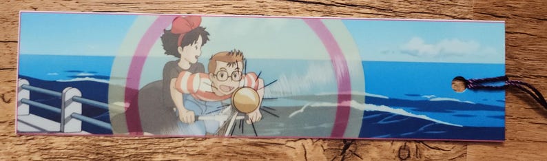 Peut inclure: Un marque-page rectangulaire pr&eacute;sentant une illustration color&eacute;e de style anime de deux personnages faisant du v&eacute;lo ensemble. La sc&egrave;ne montre un arri&egrave;re-plan d'oc&eacute;an bleu avec une rambarde de jet&eacute;e &agrave; gauche et un petit trou avec une ficelle sombre &agrave; droite.