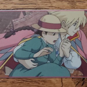 Peut inclure: Un marque-page rectangulaire en papier illustr&eacute; de deux personnages anim&eacute;s dans un d&eacute;cor urbain. L'un porte un chapeau de paille et une robe bleu sarcelle, l'autre a des cheveux blonds et une cape rose. Un cordon rose est fix&eacute; sur le c&ocirc;t&eacute; gauche.