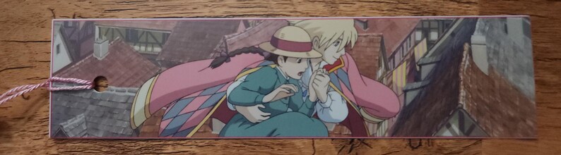 Peut inclure: Un marque-page rectangulaire en papier illustr&eacute; de deux personnages anim&eacute;s dans un d&eacute;cor urbain. L'un porte un chapeau de paille et une robe bleu sarcelle, l'autre a des cheveux blonds et une cape rose. Un cordon rose est fix&eacute; sur le c&ocirc;t&eacute; gauche.