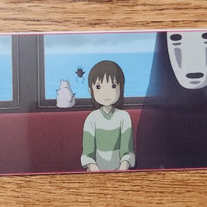 Peut inclure: Un marque-page rectangulaire en papier illustr&eacute; d'une sc&egrave;ne du Voyage de Chihiro avec Chihiro et Sans-Visage assis dans un train. L'image montre une vue sur l'oc&eacute;an bleu par les fen&ecirc;tres, une petite cr&eacute;ature blanche et un cordon noir &agrave; gauche.