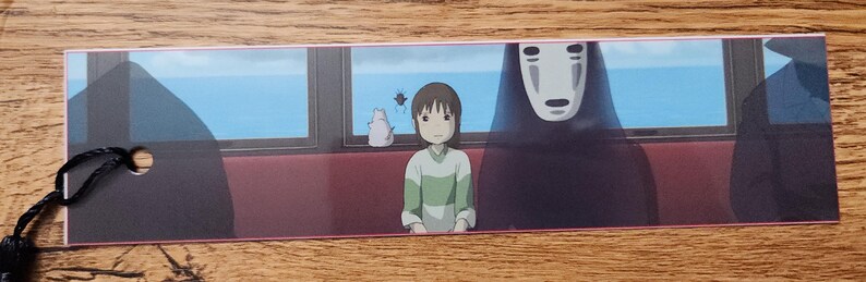Peut inclure: Un marque-page rectangulaire en papier illustr&eacute; d'une sc&egrave;ne du Voyage de Chihiro avec Chihiro et Sans-Visage assis dans un train. L'image montre une vue sur l'oc&eacute;an bleu par les fen&ecirc;tres, une petite cr&eacute;ature blanche et un cordon noir &agrave; gauche.
