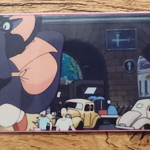 Peut inclure: Un marque-page rectangulaire en papier pr&eacute;sentant une illustration color&eacute;e de style anime avec un personnage aux cheveux sombres et un chat noir, sur fond de rue urbaine avec des voitures anciennes, dot&eacute; d'un trou et d'un cordon rouge.
