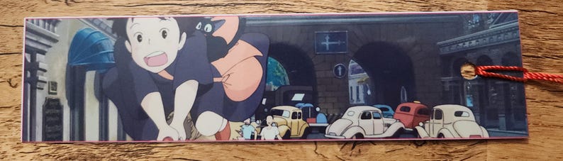 Peut inclure: Un marque-page rectangulaire en papier pr&eacute;sentant une illustration color&eacute;e de style anime avec un personnage aux cheveux sombres et un chat noir, sur fond de rue urbaine avec des voitures anciennes, dot&eacute; d'un trou et d'un cordon rouge.
