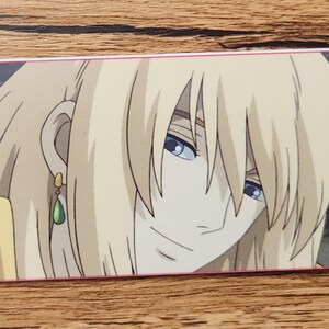 Peut inclure: Un marque-page rectangulaire en papier pr&eacute;sentant une illustration imprim&eacute;e d'un personnage blond aux yeux bleus portant une boucle d'oreille en forme de goutte verte. Le personnage est devant un fond sombre, avec un trou et un cordon jaune.