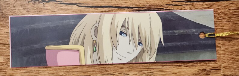 Peut inclure: Un marque-page rectangulaire en papier pr&eacute;sentant une illustration imprim&eacute;e d'un personnage blond aux yeux bleus portant une boucle d'oreille en forme de goutte verte. Le personnage est devant un fond sombre, avec un trou et un cordon jaune.