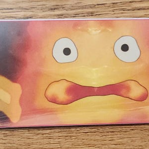Peut inclure: Un marque-page rectangulaire en papier pr&eacute;sentant une illustration graphique d'un personnage de flamme orange et jaune stylis&eacute; avec de grands yeux et une bouche ouverte, flanqu&eacute; de deux petites figurines en pierre, avec un pompon rouge.