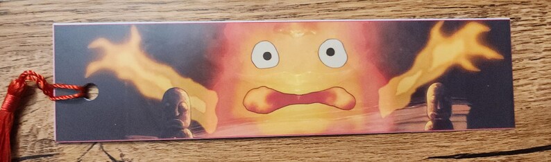 Peut inclure: Un marque-page rectangulaire en papier pr&eacute;sentant une illustration graphique d'un personnage de flamme orange et jaune stylis&eacute; avec de grands yeux et une bouche ouverte, flanqu&eacute; de deux petites figurines en pierre, avec un pompon rouge.