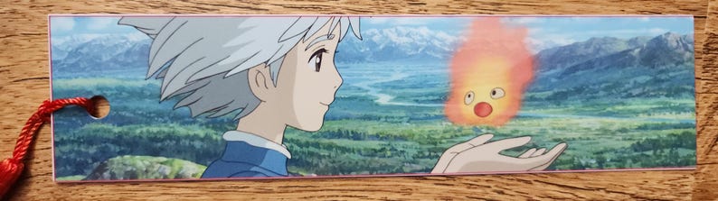Peut inclure: Un marque-page rectangulaire en papier illustr&eacute; d'un personnage de style anime aux cheveux blancs courts regardant un petit esprit de feu orange brillant flottant au-dessus d'une paume ouverte, devant des collines et des montagnes lointaines.