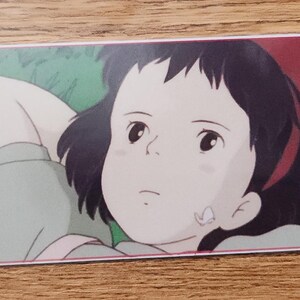 Peut inclure: Un marque-page rectangulaire en papier avec une illustration imprim&eacute;e d'une personne aux cheveux fonc&eacute;s allong&eacute;e dans l'herbe verte. La personne a les mains derri&egrave;re la t&ecirc;te et regarde vers le haut. Une petite radio rouge est pos&eacute;e dans l'herbe. Une ficelle rouge et blanche est attach&eacute;e.