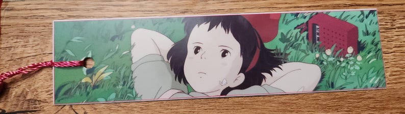 Peut inclure: Un marque-page rectangulaire en papier avec une illustration imprim&eacute;e d'une personne aux cheveux fonc&eacute;s allong&eacute;e dans l'herbe verte. La personne a les mains derri&egrave;re la t&ecirc;te et regarde vers le haut. Une petite radio rouge est pos&eacute;e dans l'herbe. Une ficelle rouge et blanche est attach&eacute;e.